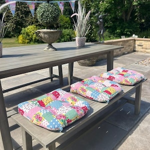May include: Three patchwork cushions in various colours sit on a grey wooden bench. The cushions have floral and polka dot patterns in pink, blue, yellow, and green. The bench is part of a grey wooden outdoor dining set.