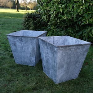 May include: Two weathered, square metal planters with a tree design on the front. The planters are grey and sit on green grass. The background includes trees and a green hedge.