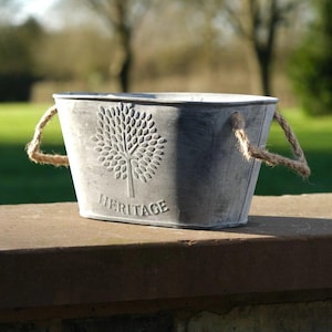 May include: A grey metal planter with rope handles. The planter has an embossed tree design and the word "HERITAGE". It sits on a brick and stone surface, with a blurred green background.