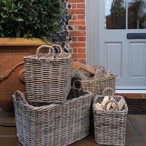 May include: A collection of four grey wicker log baskets of different sizes, some containing firewood. The baskets, with woven handles, are positioned on a stone patio near a brick wall and a front door.