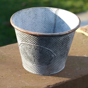 May include: A small, galvanised metal bucket with a textured pattern. The bucket has a brown rim and a decorative oval emblem with a tree design. The word "HERITAGE" is printed on the emblem. The bucket is sitting on a brown surface.