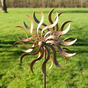 May include: A copper-toned metal garden ornament with a sunburst design. The outdoor art piece features curved, swirling blades radiating from a central sphere, mounted on a slender pole. The sculpture is set against a backdrop of green grass and trees.