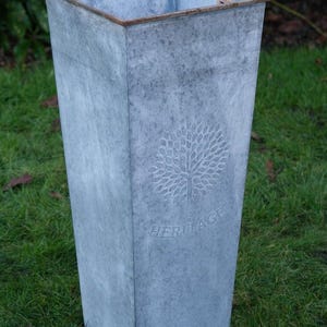 May include: A tall, rectangular galvanised metal planter with a tree design and the word "HERITAGE" embossed on the front. The planter has a slightly tapered shape and a weathered, grey finish. The top edge has a brown trim.