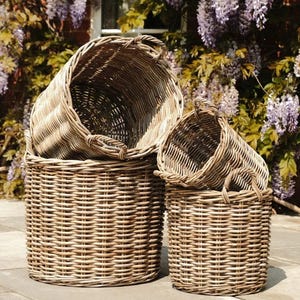 May include: Four woven wicker baskets of varying sizes are stacked together. The baskets are cylindrical with rounded handles. The natural-toned baskets are set on a stone patio, with purple wisteria flowers in the background.