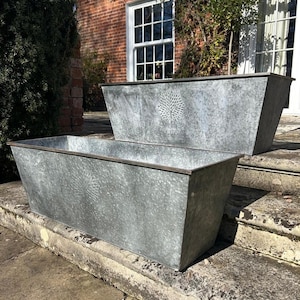 May include: Two rectangular galvanized metal planters. Each planter has a tree design and the word "HERITAGE" etched on the front. The planters are on a stone surface, with a brick building and greenery in the background.