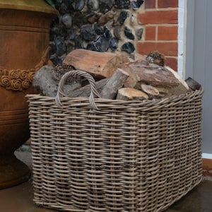 May include: A rectangular woven wicker basket with two integrated handles, filled with a collection of natural brown firewood logs. The basket features a tight, horizontal weave pattern and is constructed from light brown, weathered willow branches.