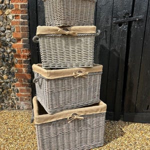 May include: Four grey wicker storage baskets, stacked. Each basket has a tan fabric lining and a decorative bow. The baskets have rope handles and are of varying sizes. Ideal for storage.