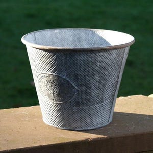 May include: A galvanized metal bucket with a textured surface and an oval emblem. The bucket has a tapered shape and a dark rim. The emblem features a tree design and the word "HERITAGE".
