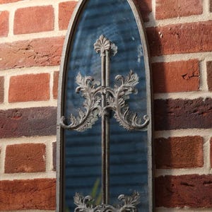 May include: A tall, arched wall mirror with a weathered grey metal frame. The mirror features a central vertical bar and two symmetrical, ornate metal scrollwork overlays with leaf-like detailing positioned in the upper and lower sections.