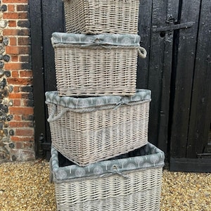 May include: Four grey wicker storage baskets of different sizes are stacked. Each basket has a fabric lining with a white tree pattern and rope handles. The baskets are against a black wooden door and a brick wall.