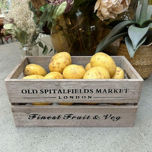 May include: A wooden crate filled with yellow potatoes. The crate is branded with "OLD SPITALFIELDS MARKET LONDON" and "Finest Fruit & Veg". The crate is made of weathered wood and is on a light-colored surface. The background includes flowers.