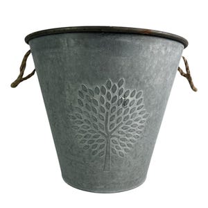 May include: A grey galvanised metal bucket featuring a tree design. The bucket has a dark brown rim and rope handles. The tree design is embossed on the front of the bucket.