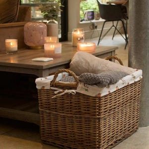 May include: A large, woven wicker basket containing folded towels, with a patterned fabric lining. Several lit candles in glass jars and a decorative vase are on a wooden coffee table. The room is bright and airy.