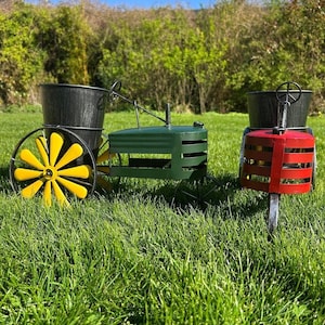 May include: Three decorative metal tractor planters in a grassy garden. One is green with a yellow wheel, another is red, and the third is green. Each has a black bucket for planting. The tractors are designed to be garden decor.