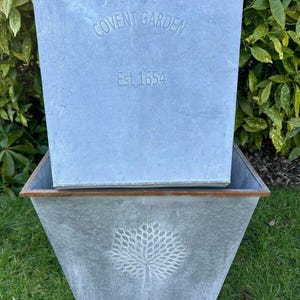 May include: Two galvanized metal planters. The top planter has the text "COVENT GARDEN Est 1654". The bottom planter features a tree design. Both planters have a brown trim and are set on green grass.