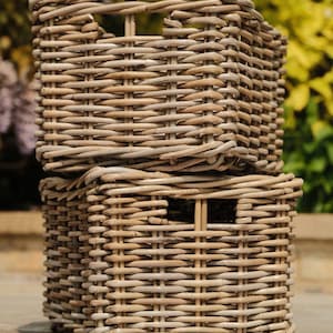 May include: Two stacked rectangular wicker storage baskets. The baskets are woven from light brown, natural-looking material. Each basket has a rectangular opening on the front and a handle-like opening at the top. The background is blurred with green and yellow foliage.