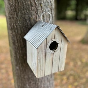 May include: A wooden birdhouse with a corrugated metal roof and a circular entry hole. The birdhouse is mounted on a tree trunk and has a metal loop for hanging. The wood has a weathered, light-coloured finish.