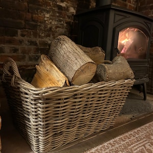 May include: A light grey wicker basket, full of logs, sits beside a fireplace. The basket has handles and is rectangular. The logs are various sizes and shapes. A black stove with a fire burning inside is in the background.