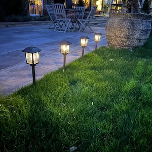 May include: A garden scene at dusk, showing four solar-powered lights with a warm, inviting glow, positioned in a line on a green lawn. The lights have a traditional lantern shape with a dark metal finish. The background includes a patio, garden furniture, and a large plant pot.