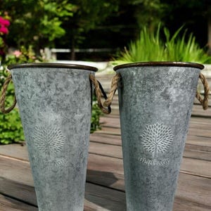 May include: Two galvanised metal planters with rope handles. Each planter has a tree design and the word "HERITAGE" embossed on the front. The planters are grey with a brown rim and are on a wooden deck.