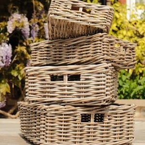 May include: Four rectangular wicker storage baskets, stacked. The baskets are a natural light brown colour and have cut-out handles. They are arranged on a stone surface, with a backdrop of foliage and a brick building. The baskets are made of woven material.