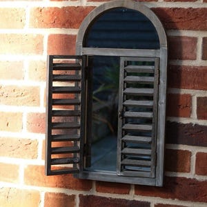 May include: A wooden window mirror with an arched top and shutter doors. The shutters are open, showing the mirror and a glimpse of the background. The frame has a weathered, grey finish, and is mounted on a red brick wall.