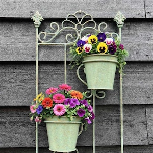 May include: A wall-mounted planter featuring two light green buckets overflowing with vibrant flowers. The metal frame has an ornate design and is fixed to a grey wooden wall. The flowers include pink, orange, purple, yellow, and blue blooms.