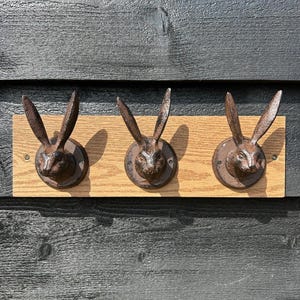 May include: Three bronze-coloured metal rabbit head hooks mounted on a light brown wooden board. The hooks feature detailed rabbit faces and long ears. The board is attached to a dark wooden surface.