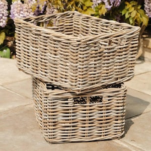 May include: Two rectangular, light brown wicker baskets are stacked. The top basket is larger than the bottom one. Both have a woven design. The background shows purple flowers and green foliage. The baskets are empty and sit on a stone surface.