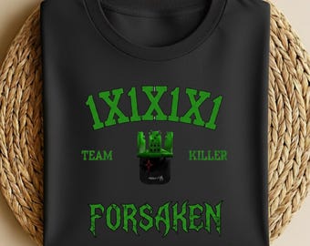 May include: Black t-shirt with green text and graphics. The text includes "1X1X1X1", "TEAM KILLER", and "FORSAKEN". A green graphic of a castle is centered above the word "FORSAKEN". The shirt is folded on a woven surface.