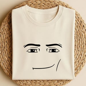 May include: Cream-colored t-shirt featuring a minimalist black line drawing of a face. The design includes eyebrows, eyes, and a smiling mouth. The shirt is folded and placed on a woven, circular mat.