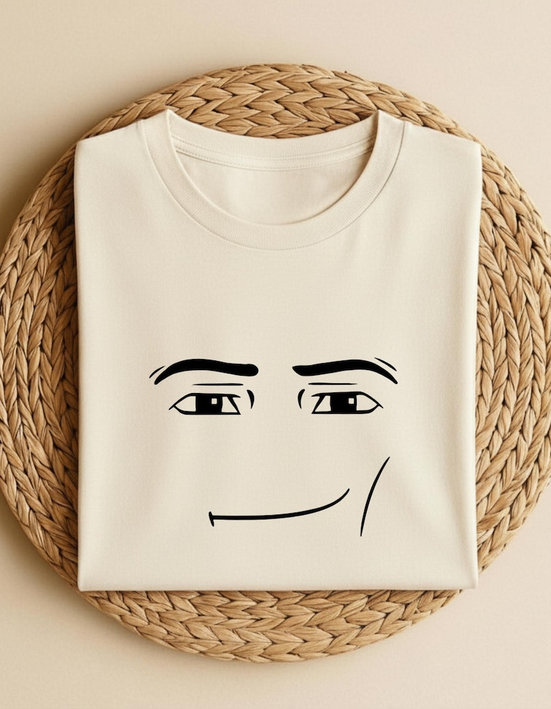 May include: Cream-colored t-shirt featuring a minimalist black line drawing of a face. The design includes eyebrows, eyes, and a smiling mouth. The shirt is folded and placed on a woven, circular mat.