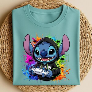 May include: A light teal t-shirt featuring a cartoon character with large ears, holding a video game controller. The character is blue with a black hoodie and the word "CHAIHA" on the hoodie. Colorful paint splatters surround the character.