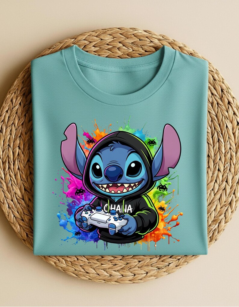 May include: A light teal t-shirt featuring a cartoon character with large ears, holding a video game controller. The character is blue with a black hoodie and the word "CHAIHA" on the hoodie. Colorful paint splatters surround the character.