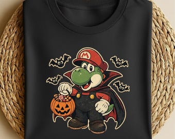 Może przedstawiać: Czarny t-shirt z grafiką przedstawiającą zielonego dinozaura w czerwonej czapce z logo M i pelerynie wampira. Postać trzyma pomarańczowe wiaderko w kształcie dyni wypełnione słodyczami, w otoczeniu czterech małych czarnych nietoperzy.