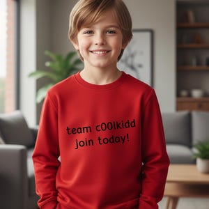 May include: A young person wearing a red sweatshirt with the text "team c00lkidd join today!" in black. The sweatshirt is a solid red color. The person is smiling and looking at the camera.