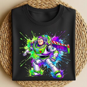 K&ouml;nnte beinhalten: Schwarzes T-Shirt mit einer farbenfrohen Grafik von Buzz Lightyear aus Toy Story. Das Design zeigt Buzz in seinem Raumanzug mit einem lebendigen Splash-Art-Hintergrund in Gr&uuml;n-, Lila-, Blau- und Rosat&ouml;nen. Das T-Shirt ist sauber gefaltet.