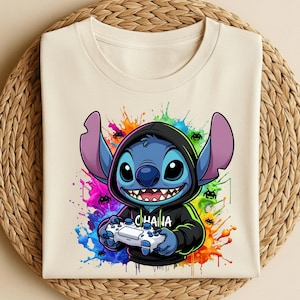 May include: Cream-colored t-shirt featuring a blue cartoon character in a black hoodie, holding a white game controller. The design includes the word "CHANA" and colorful paint splatters. The shirt is folded on a woven mat.