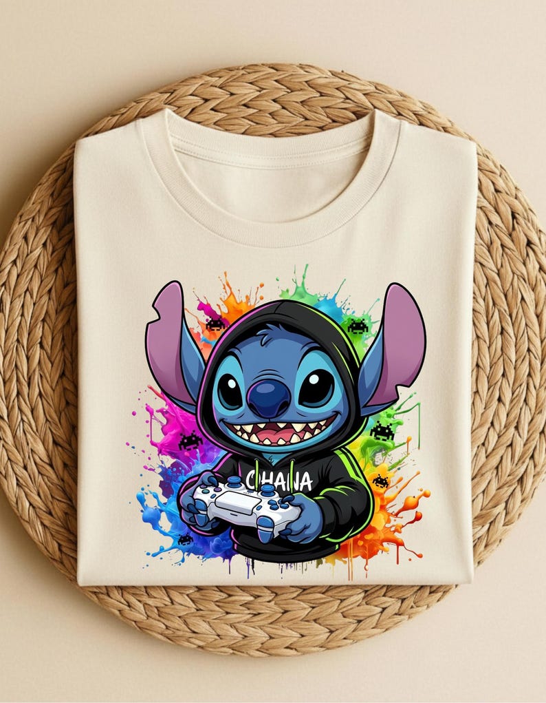May include: Cream-colored t-shirt featuring a blue cartoon character in a black hoodie, holding a white game controller. The design includes the word "CHANA" and colorful paint splatters. The shirt is folded on a woven mat.