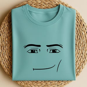 May include: A light teal t-shirt featuring a black cartoon face with a smiling expression. The shirt is folded and placed on a woven, circular placemat. The design includes eyebrows, eyes, and a curved line for the mouth.