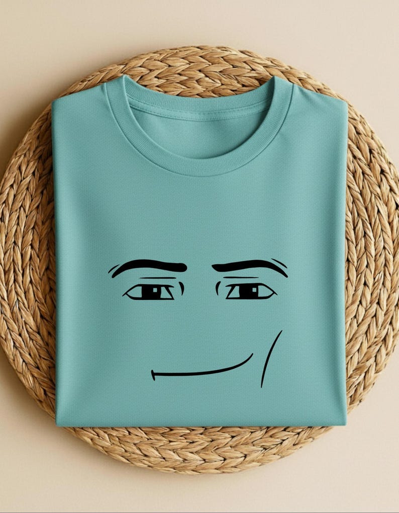 May include: A light teal t-shirt featuring a black cartoon face with a smiling expression. The shirt is folded and placed on a woven, circular placemat. The design includes eyebrows, eyes, and a curved line for the mouth.