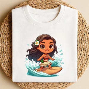 May include: White crew neck t-shirt featuring a cartoon illustration of a girl surfing. The girl has brown hair, a flower, and is wearing a skirt. The design includes blue water and a surfboard.