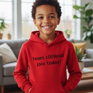 May include: A smiling child wearing a red hoodie with a front pocket. The hoodie has the text "team c00lkidd join today!" printed in black. The child is standing in front of a window and a gray couch.