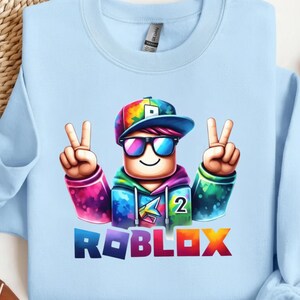 Puede incluir: Sudadera azul claro con un personaje colorido de Roblox con gafas de sol, un signo de la paz y la palabra "ROBLOX" en letras vibrantes. El personaje lleva una gorra arco&iacute;ris y una sudadera con capucha. Tambi&eacute;n es visible el n&uacute;mero "2".