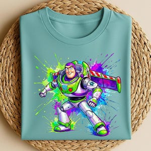 K&ouml;nnte beinhalten: Ein hellt&uuml;rkisfarbenes T-Shirt mit einer farbenfrohen Grafik von Buzz Lightyear. Die Zeichentrickfigur ist in ihrem Raumanzug vor einem lebendigen Splash-Art-Hintergrund in Gr&uuml;n, Lila und Blau dargestellt. Das T-Shirt ist auf einer gewebten Matte gefaltet.