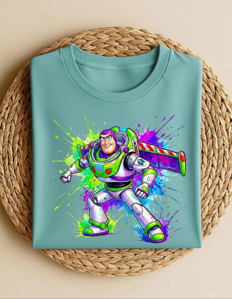 K&ouml;nnte beinhalten: Ein hellt&uuml;rkisfarbenes T-Shirt mit einer farbenfrohen Grafik von Buzz Lightyear. Die Zeichentrickfigur ist in ihrem Raumanzug vor einem lebendigen Splash-Art-Hintergrund in Gr&uuml;n, Lila und Blau dargestellt. Das T-Shirt ist auf einer gewebten Matte gefaltet.
