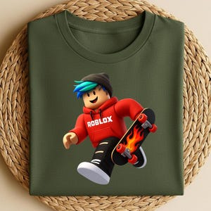 May include: Olive green t-shirt featuring a Roblox character in a red hoodie with the word "ROBLOX" printed on it. The character is skateboarding on a black skateboard with flames. The character has blue and green hair and is wearing a black beanie.