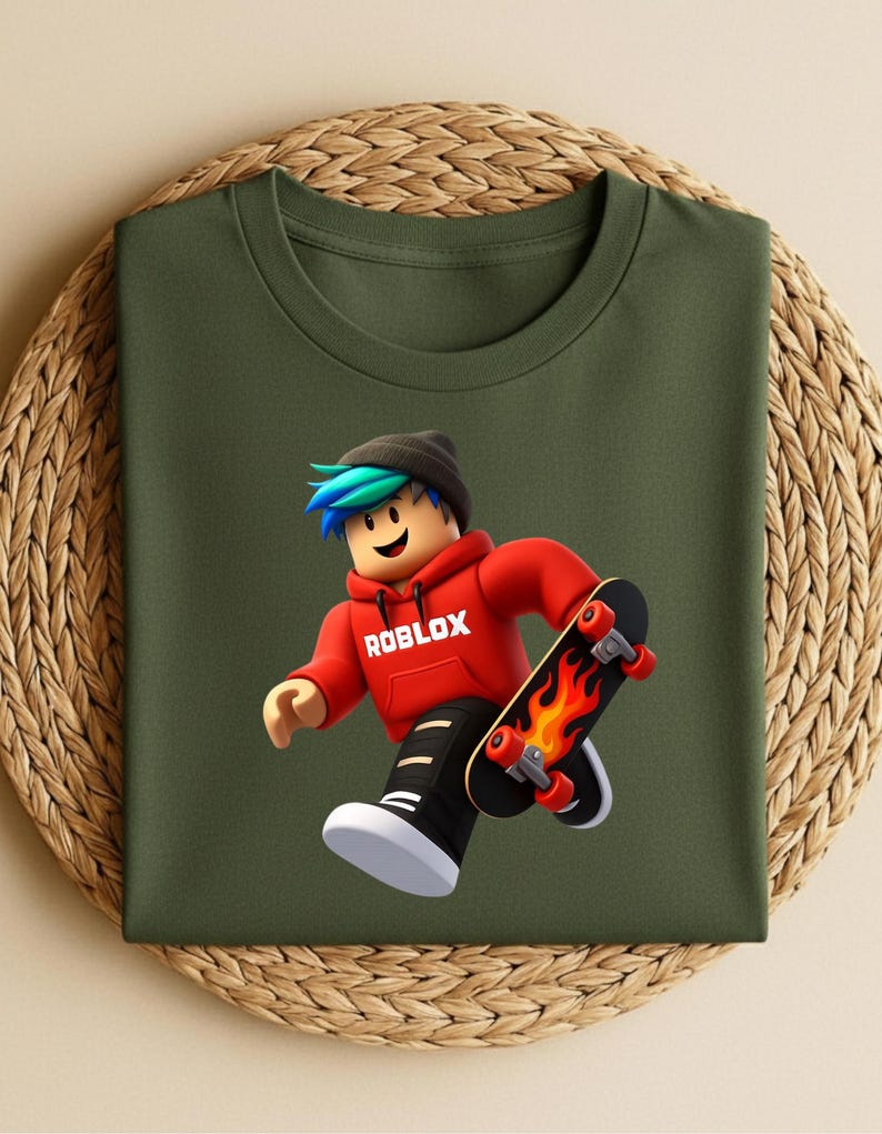 May include: Olive green t-shirt featuring a Roblox character in a red hoodie with the word "ROBLOX" printed on it. The character is skateboarding on a black skateboard with flames. The character has blue and green hair and is wearing a black beanie.