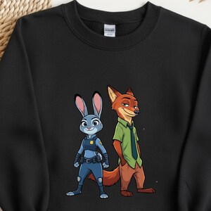 以下が含まれることがあります： 黒いクルーネックのスウェットシャツ。青いウサギのジュディ・ホップスと赤いキツネのニック・ワイルドの漫画イラストが描かれています。ジュディは警察の制服を着ており、ニックは緑のシャツとネクタイを着ています。スウェットシャツは柔らかい素材で作られています。
