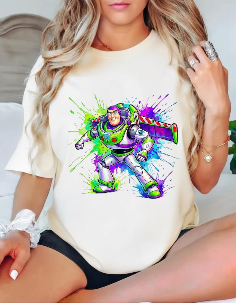 K&ouml;nnte beinhalten: Cremefarbenes T-Shirt mit einer farbenfrohen Grafik von Buzz Lightyear aus Toy Story. Die Figur ist in ihrem Raumanzug dargestellt, umgeben von lebendigen Farbspritzern in Gr&uuml;n, Lila und Blau. Das Design ist auf dem Shirt zentriert.
